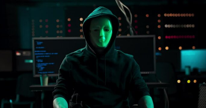 Wanted Hacker with Masked Face looks into the Camera. In the Background His Operating Room with Multiple Displays and Cables. Anonymous man covers her mouth with her finger in a gesture of silence
