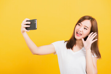 Naklejka premium Asian happy portrait beautiful cute young woman teen smiling standing making selfie photo, video call on smartphone raise hand to say hello isolated, studio shot yellow background with copy space