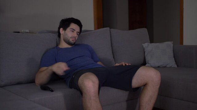 A bored and tired young man is sitting on the sofa in the living room and watching TV. Not finding an interesting TV channel, the person turns off the TV and leaves