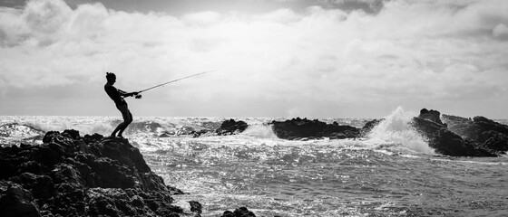 A fisherman fishing on the ocean sea with his pole, standing on the shore rocks with waves.