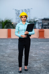 Full length portrait of beautiful female engineer wearing a protective helmet and holding files in hand while looking at camera.