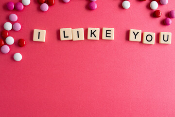 the words i like you written as a flat lay in wood scrabble tiles on a plain red background surrounded by red, white, and pink candies