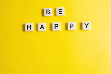 the words be happy written as a flat lay in wood scrabble tiles on a plain yellow background