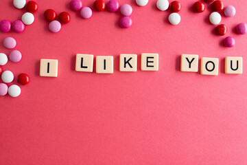 the words i like you written as a flat lay in wood scrabble tiles on a plain red background surrounded by red, white, and pink candies