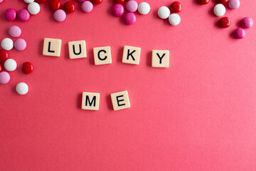 the words lucky me written as a flat lay in wood scrabble tiles on a plain red background surrounded by red, white, and pink candies