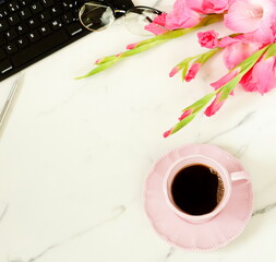 Flat lay, top view women's office desk with  flowers and cup of coffee. Holiday workspace with laptop, pink gladioluses ,accessories, glasses, cup of coffee on marble desk  background.  Copy space
