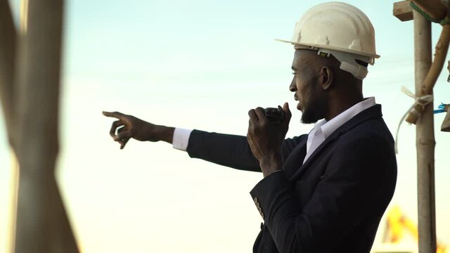 african american architect in suit and hard hat talking by walkie talkie control and communication with worker outdoors on construction site. contractor using radio operation in industry