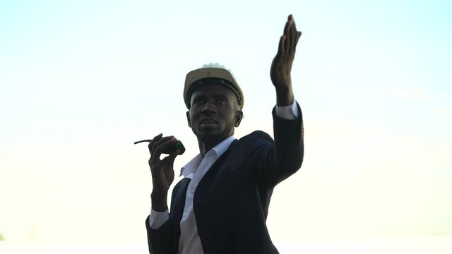 african american architect in suit and hard hat talking by walkie talkie control and communication with worker outdoors on construction site. contractor using radio operation in industry