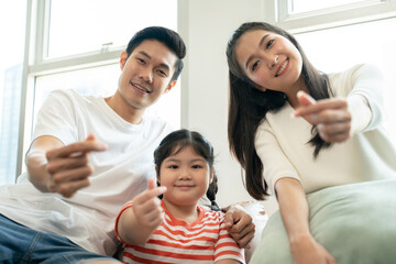 Happy Asian family sitting on sofa and showing hand sign of mini heart,looking at camera to video call,family relaxed activity.Blogger family sharing good positive mood for viewers.