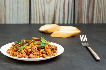 Mexican dish in a white plate on a black table against a background of a wooden wall. White bread and a fork.