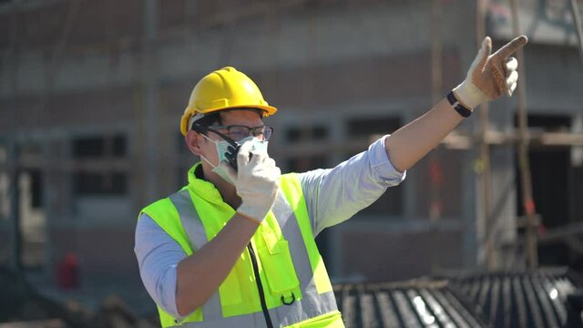 asian builder in hard hat wearing protective face mask talking by walkie talkie control and communication with worker outdoors on construction site. contractor using radio operation in industry