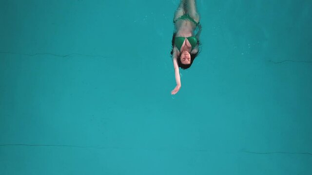 Woman in a green bikini swimming backward in a pool aerial