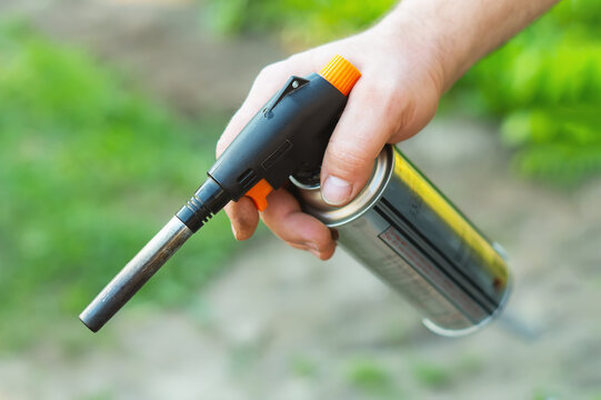 A man holds a gas burner in his hands close-up against the background of green grass. Use of a gas burner with a cylinder to start a fire.