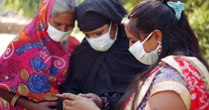 Close-up of group of women in ethnic clothes, Muslim Hijab and Hindu Sari with face mask discuss share using smart phone mobile technology communication device  during coronavirus covid-19 lockdown