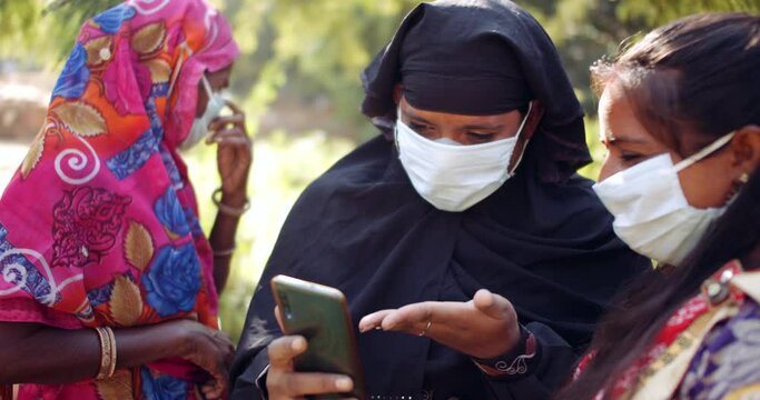 Close-up of group of women in ethnic clothes, Muslim Hijab and Hindu Sari with face mask discuss share using smart phone mobile technology communication device  during coronavirus covid-19 lockdown