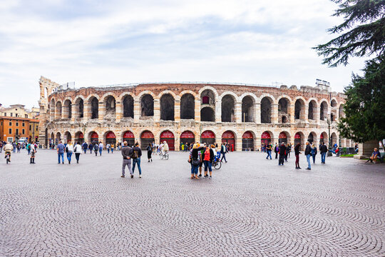Tourists walking around the Piazza Bra square on a cloudy day, inspect the Arena and take pictures against its background in Verona, Italy.