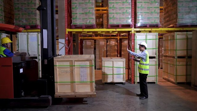 Asian male foreman checking goods inventory stock and working with warehouse worker in distribution fulfillment center. Labor man move cardboard box on forklift truck on the shelf in retail warehouse.