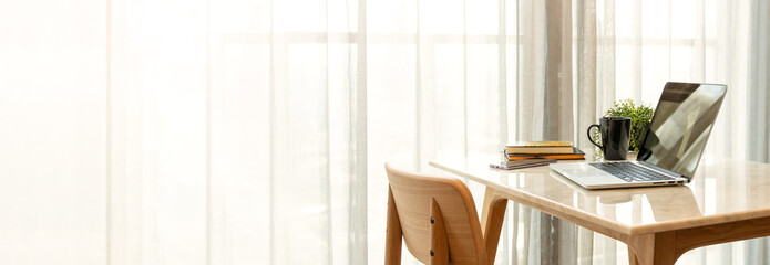 Beautiful workspace - online remote work from home concept. A wooden table with computer laptop, smartphone, coffee cup beside the window balcony with sun light shine through the curtain. Banner