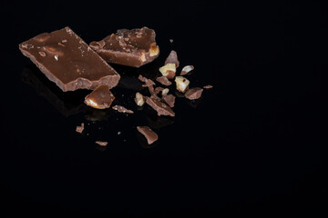 Pieces of milk chocolate with crushed hazelnuts on reflective black background. Free space for text.