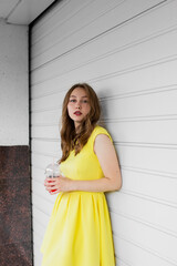 Fototapeta premium Young woman in illuminating yellow dress trend color of 2021 standing in front of white wall, holding glass of lemonade and looking at camera. Girl having fun. Urban style and fashion. Casual outfit