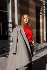 Portrait of a young woman taking off coat, looking at camera and standing on street in front of window and brown wall. Caucasian girl having fun outside. Urban style and fashion. Casual outfit