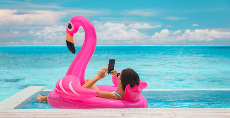 Relaxing woman floating in flamingo inflatable swimming pool toy at luxury resort using mobile...