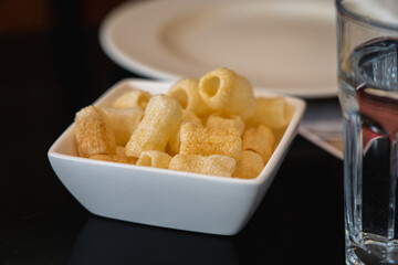 Fryums served in a small white bowl on a black table. A glass of water in the foreground and a white plate in the background.