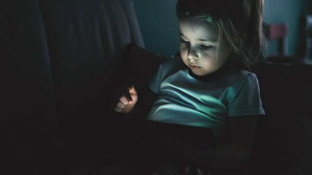 Little preschool girl use digital tablet technology device lying at sofa in dark room. Eyes care, gadget addiction concept. Cinematic noise.