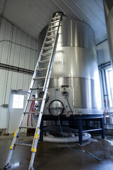 A Large Stainless Steel Commercial Wine Holding Fermentation Blending Tank with a Ladder Propped Up Against It, Cement Ground, Indoors - Oregon