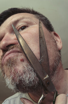 An adult man cuts his own beard with old huge scissors.