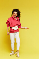 Young woman in a pink hoodie, white pants and sunglasses posing over yellow background. Model in sportswear with beltbag on the yellow background