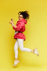 Fototapeta na wymiar Positive model girl in casual sportswear and sunglasses jumping over a yellow background. Young beautiful woman in a pink hoodie and white pants going crazy