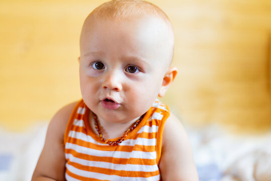 A portrait of a cute adorable curious  baby boy looking at the camera wearing amber teething baby necklace in a sleeveless striped orange top. Trying to say something, lips folded tube.