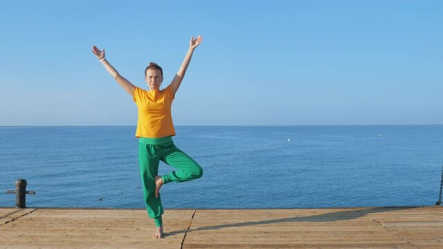 A young woman in colorful clothes practices yoga by the sea or ocean. Training tree pose. Self-knowledge and healthy lifestyle concept.