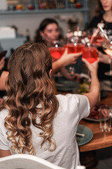 The bride sits at the table from behind and holds cocktails in her hands with her friends. In the background are figures of girls.