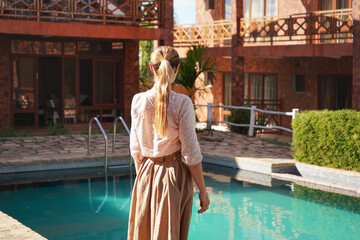 Young woman in summer dress and skirt looking into empty hotel resort pool, view from behind