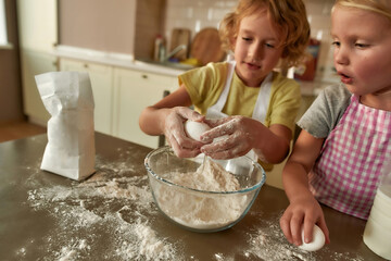 Little helpers. Two kids, boy and girl adding eggs and other ingredients while cooking, preparing...