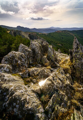 Landscape of rocks with green plants, dramatic sky with clouds and distant mountains.
