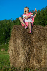 Naklejka na ściany i meble Blonde woman in bright pink suit and white T-shirt sits on haystack against background of mountains, trees and round rolls of hay with phone and takes selfie with hand outstretched. Summer vacation