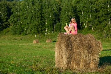 Naklejka na ściany i meble Blonde woman in bright pink suit and white T-shirt sits on haystack against background of mountains, trees and round rolls of hay with phone and takes selfie with hand outstretched. Summer vacation