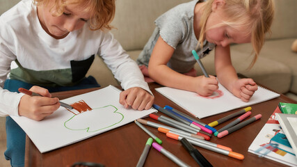 Cropped shot of adorable little children, boy and girl looking focused while drawing on paper using marker pen, sitting together on a couch at home