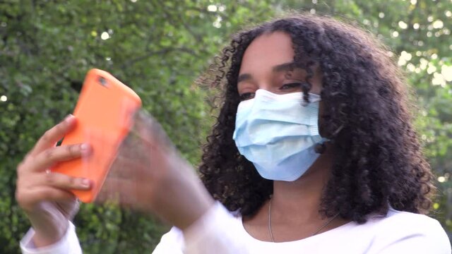 Beautiful mixed race African American girl teenager young woman wearing a face mask during COVID-19 Coronavirus pandemic using her smartphone or cell phone for social media