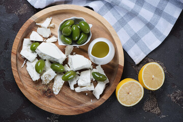 Wooden serving tray with chunks of feta, green olives, olive oil and lemon, flatlay on a brown stone background