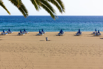 Fototapeta na wymiar Blue sunbeds and umbrellas on empty beach in Puerto del Carmen, Lanzarote. Tourism decline, pandemic crisis, economy recession concepts
