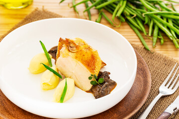 Roasted chicken breast with mashed potatoes, green beans and mushroom sauce aside a set of cutlery, olive oil and a pile natural green beans on a jute place mat oak wood table.
