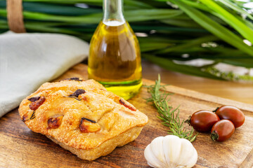 Italian focaccia with confit garlic cloves and rosemary, alongside some tomatoes,  a garlic bulb, olive oil and rosemary twig on a chopping board by a leaf background.