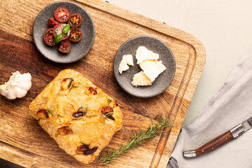 Italian focaccia with garlic cloves and rosemary, alongside tomatoes, garlic bulb, parmesan cheese and rosemary twig on a wood chopping board with knife on green tablecloth. Top view.