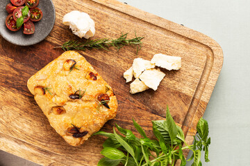 Fototapeta na wymiar Italian focaccia with garlic cloves and rosemary, alongside tomatoes, garlic bulb, parmesan cheese, rosemary and basil twig on a chopping board on green tablecloth. Top view.