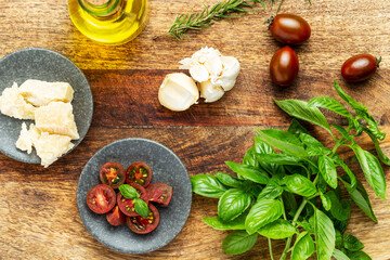 Italian cuisine ingredients: basil, tomatoes, rosemary, olive oil and parmesan cheese on a vintage wood chopping board. Flat lay, top view.