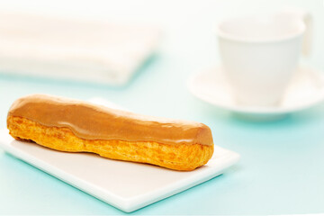 Salty caramel French eclair pastry on white plate, aside a white coffee cup and napkin over light blue background.